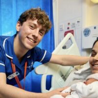 A male midwife leans over a hospital bed to care for a mum and newborn