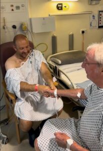 Ian Tonks (left) and Graham Sisson (right) in hospital gowns holding hands before surgery, seated beside a hospital bed 