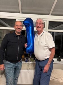 Ian Tonks (left) and Graham Sisson (right) standing indoors holding a large blue balloon shaped like the number one.