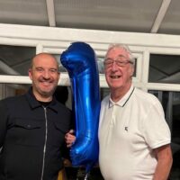 Ian Tonks (left) and Graham Sisson (right) standing indoors holding a large blue balloon shaped like the number one.