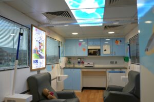 Inside the mobile cancer care unit: child-friendly space featuring colorful fish images on the walls, comfortable chairs, and a sky-themed ceiling light panel.