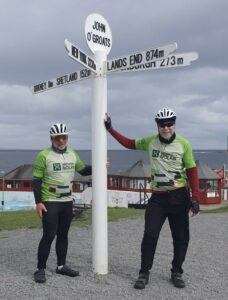 Two cyclists in matching green jerseys stand beside the John O'Groats signpost showing distances to Lands End and other locations.