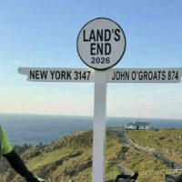 Two cyclists in helmets and matching jerseys pose with their bikes at Land's End signpost showing distances to New York and John O'Groats in a coastal landscape.