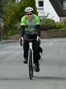 Smiling man wearing a white helmet and green cycling jersey rides a road bike on a quiet street.