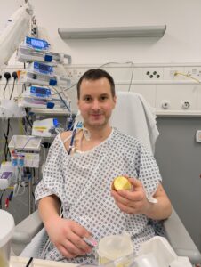 Man in hospital gown sat up in bed eating an apple.