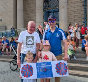 Two men and two children smiling to camera holding Team Wythenshawe signs.