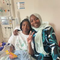 Two women smiling to camera with arms round each other, one in a hospital bed.