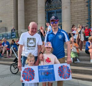 Two men and two children smiling to camera holding Team Wythenshawe signs.