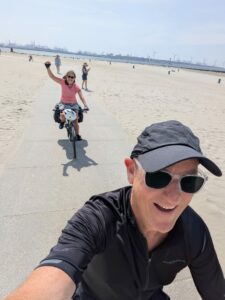 Man taking selfie while cycling bike on a beach with woman waving in the background.